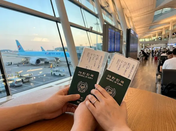 인천공항 제2터미널 출국장에서 여권과 하와이행 비행기 티켓을 들고 있는 30대 한국인 남녀의 손, 배경에는 창밖으로 비행기가 보이는 풍경