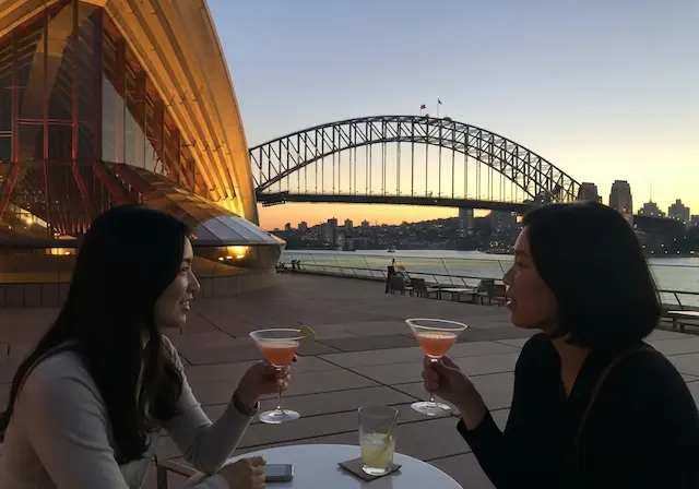 투어를 마친 후 오페라 바 야외 테라스 좌석에 앉아, 노을지는 시드니 하버 브릿지를 배경으로 칵테일 잔을 든 한국인 여성이 친구와 담소를 나누는 모습